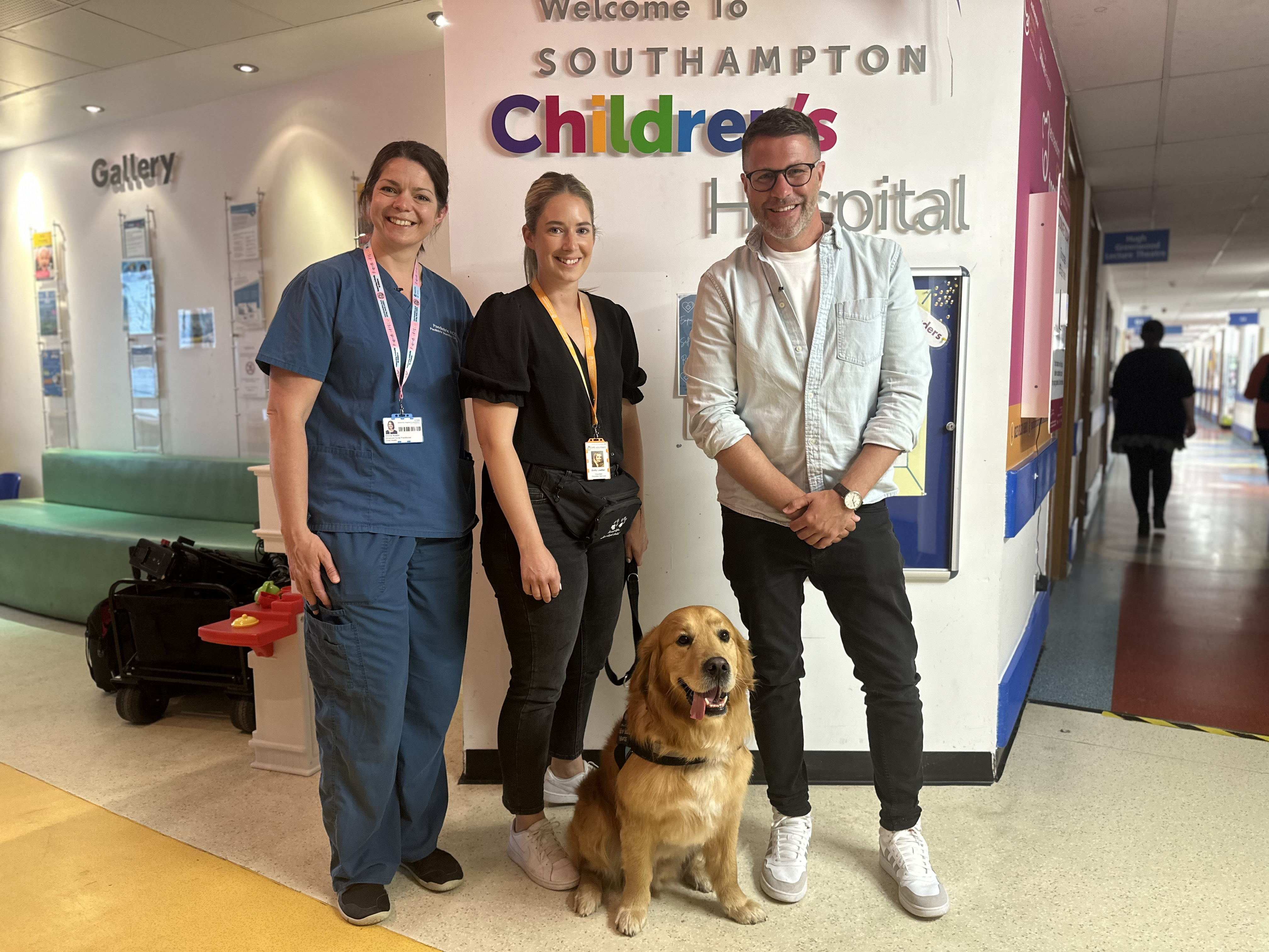 Picture shows two women and one man, with a therapy dog, outside the entrance to the children's hospital.