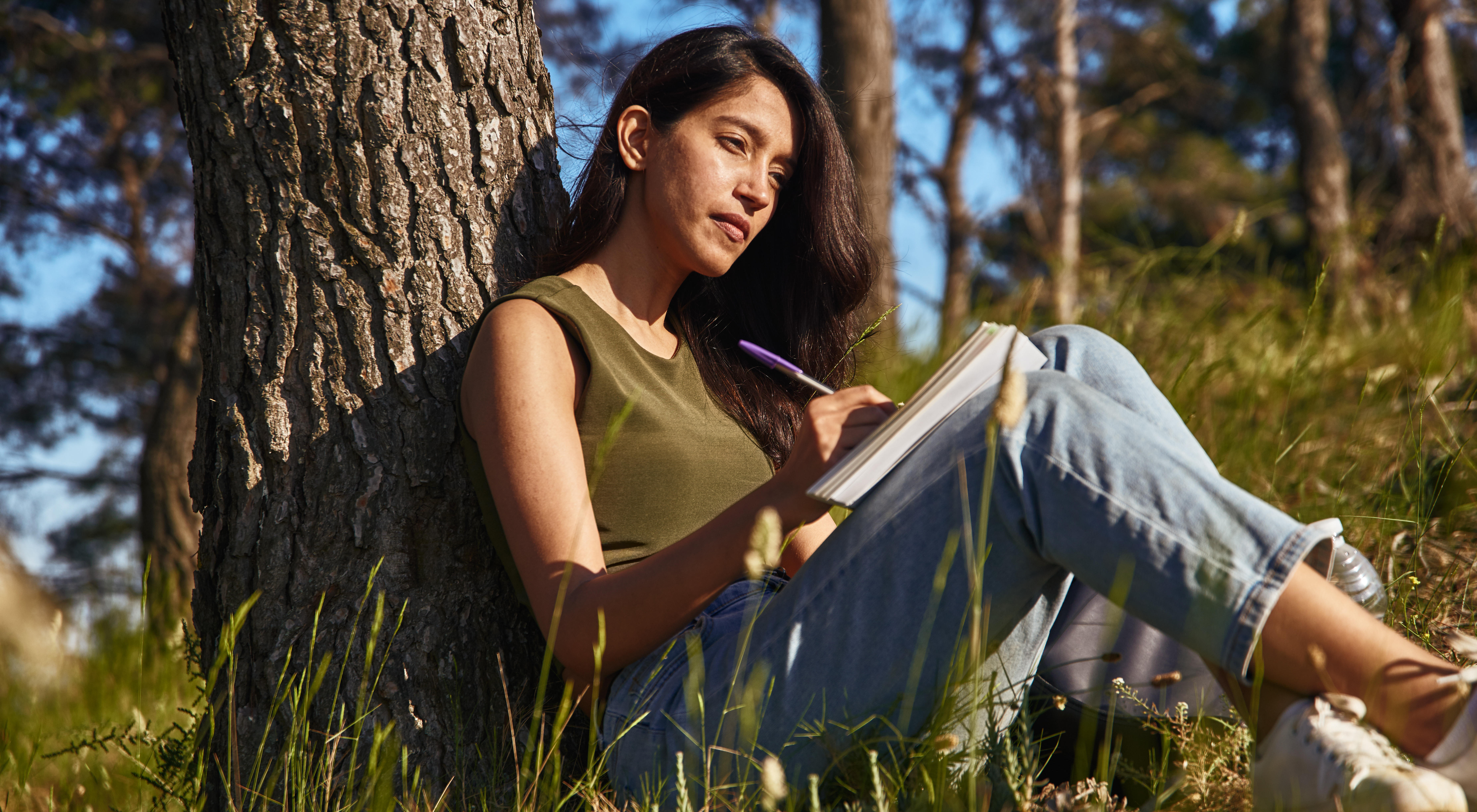 Woman sat by a tree writing on a notepad.