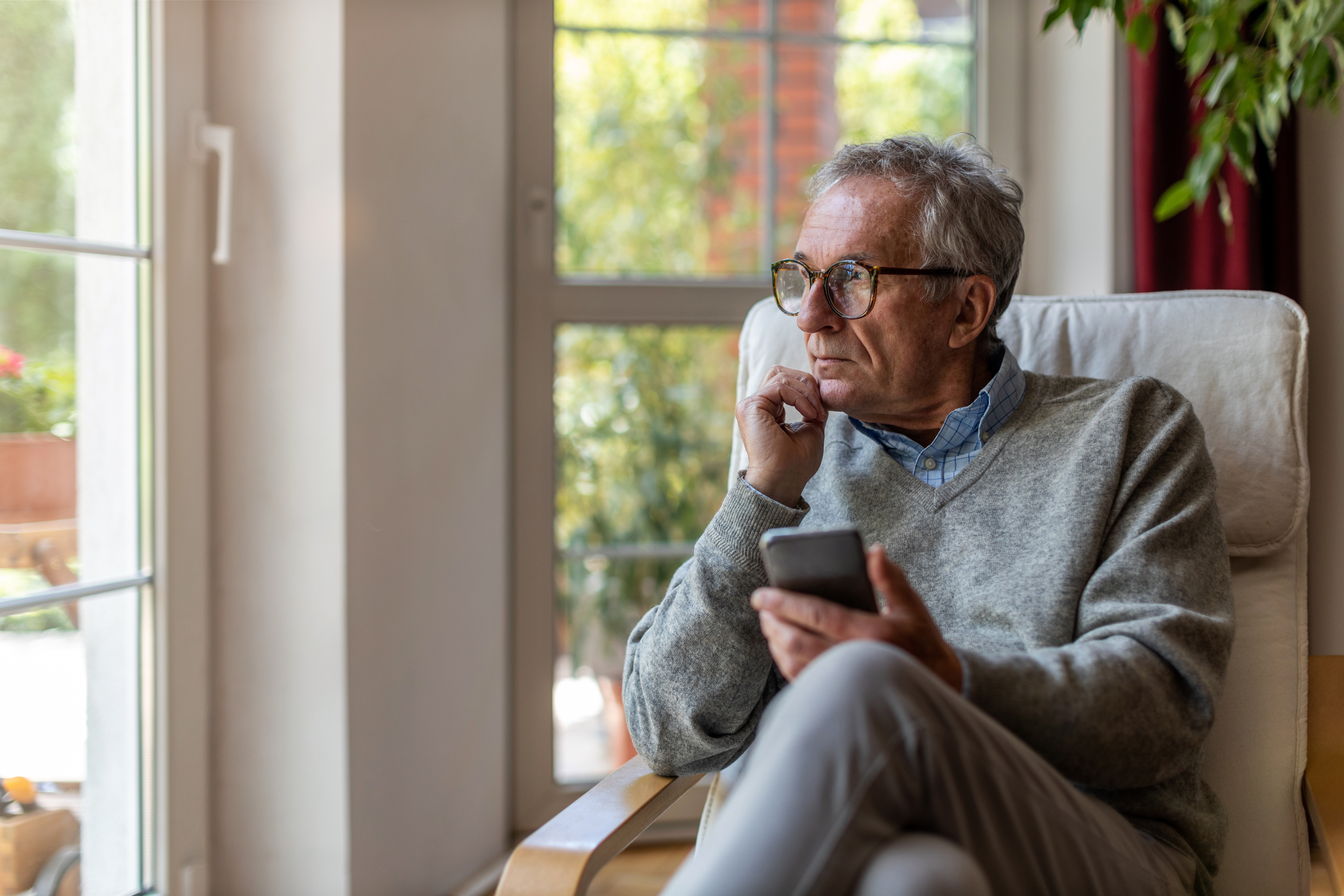 Man sitting in a chair holding his phone, gazing out of the window.